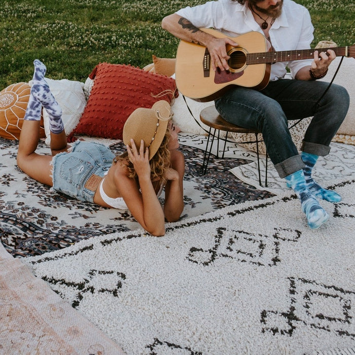 A young woman in a denim shirt and shorts lounges face-down on a patterned picnic rug wearing blue Blueberry Tie Dye Athletic socks, while a man with long hair sits in a chair playing acoustic guitar beside her in matching blue tie-dye socks. [lf]