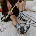 A long-haired guitarist in a brown suede jacket sits on a textured rug with his electric guitar, displaying tall Black White Tie Dye Athletic socks streaked with dramatic monochrome tie-dye swirls. [lf]
