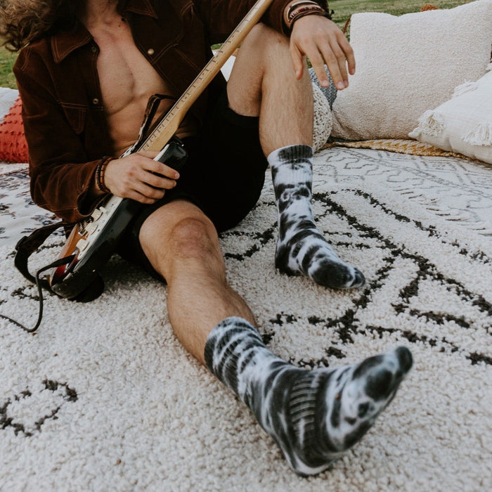 A long-haired guitarist in a brown suede jacket sits on a textured rug with his electric guitar, displaying tall Black White Tie Dye Athletic socks streaked with dramatic monochrome tie-dye swirls. [lf]