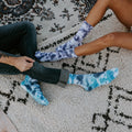 A close-up of two people sitting on a textured rug, the woman in shorts wearing purple-blue Blueberry Tie Dye Athletic socks and a man in dark jeans rolling up his cuffs over bright cyan tie-dye socks. [lf]