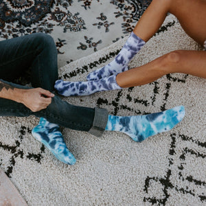 A close-up of two people sitting on a textured rug, the woman in shorts wearing purple-blue Blueberry Tie Dye Athletic socks and a man in dark jeans rolling up his cuffs over bright cyan tie-dye socks. [lf]