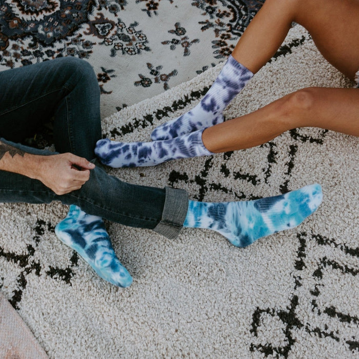 A close-up of two people sitting on a textured rug, the woman in shorts wearing purple-blue Blueberry Tie Dye Athletic socks and a man in dark jeans rolling up his cuffs over bright cyan tie-dye socks. [lf]