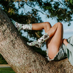 A woman in a green sweater reclines along the long arc of an oak tree branch at sunset, one leg bent and one extended in tall Black Forest Tie Dye Athletic crew socks. [lf]