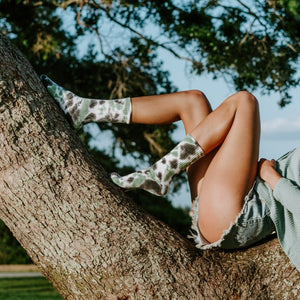 A woman in a green sweater reclines along the long arc of an oak tree branch at sunset, one leg bent and one extended in tall Black Forest Tie Dye Athletic crew socks. [lf]