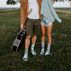 A couple walks arm in arm across a grassy field at golden hour, the man in cargo shorts holding a guitar by his side and both wearing Boot Camp Tie Dye Athletic socks in earthy tones. [lf]