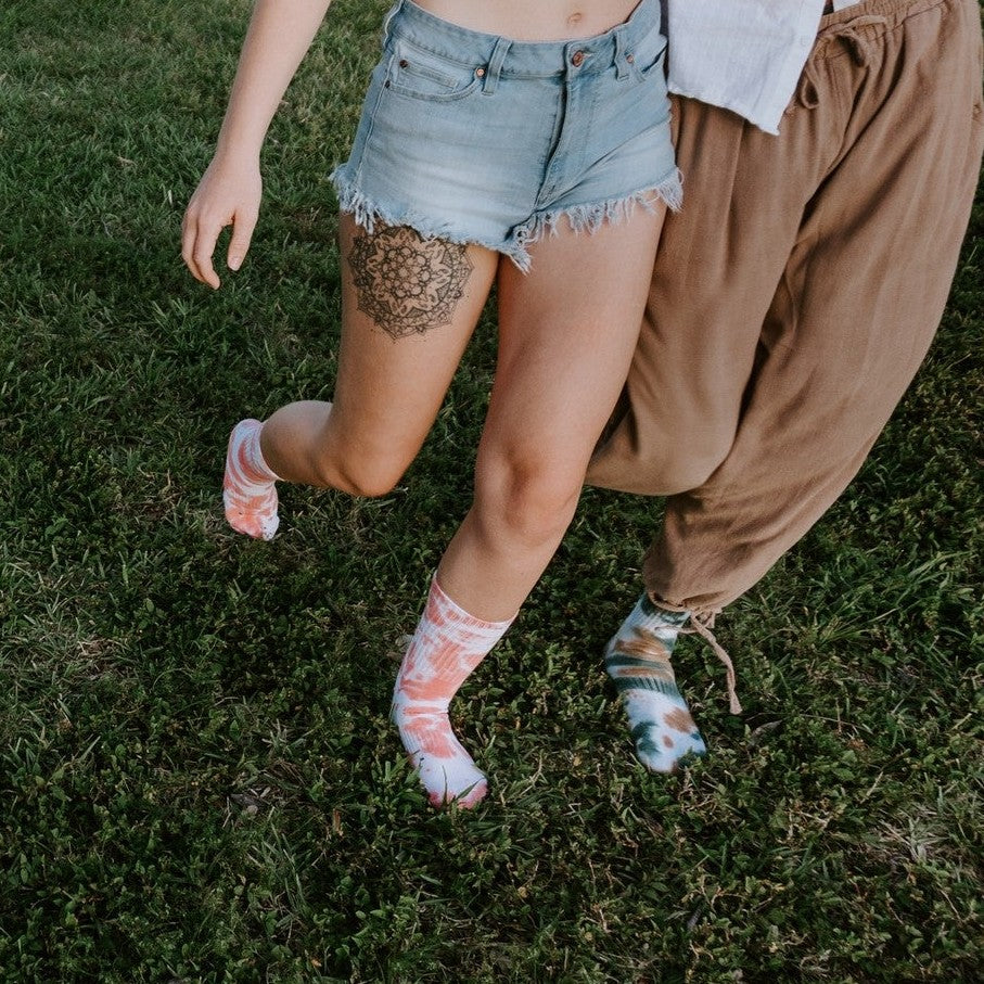 An overhead shot of friends sprawled across overlapping rugs at sunset, each person wearing a different Boot Camp Tie Dye Athletic sock pattern from teal to magenta to multicolor swirls. [lf]