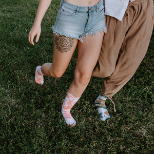 An overhead shot of friends sprawled across overlapping rugs at sunset, each person wearing a different Boot Camp Tie Dye Athletic sock pattern from teal to magenta to multicolor swirls. [lf]