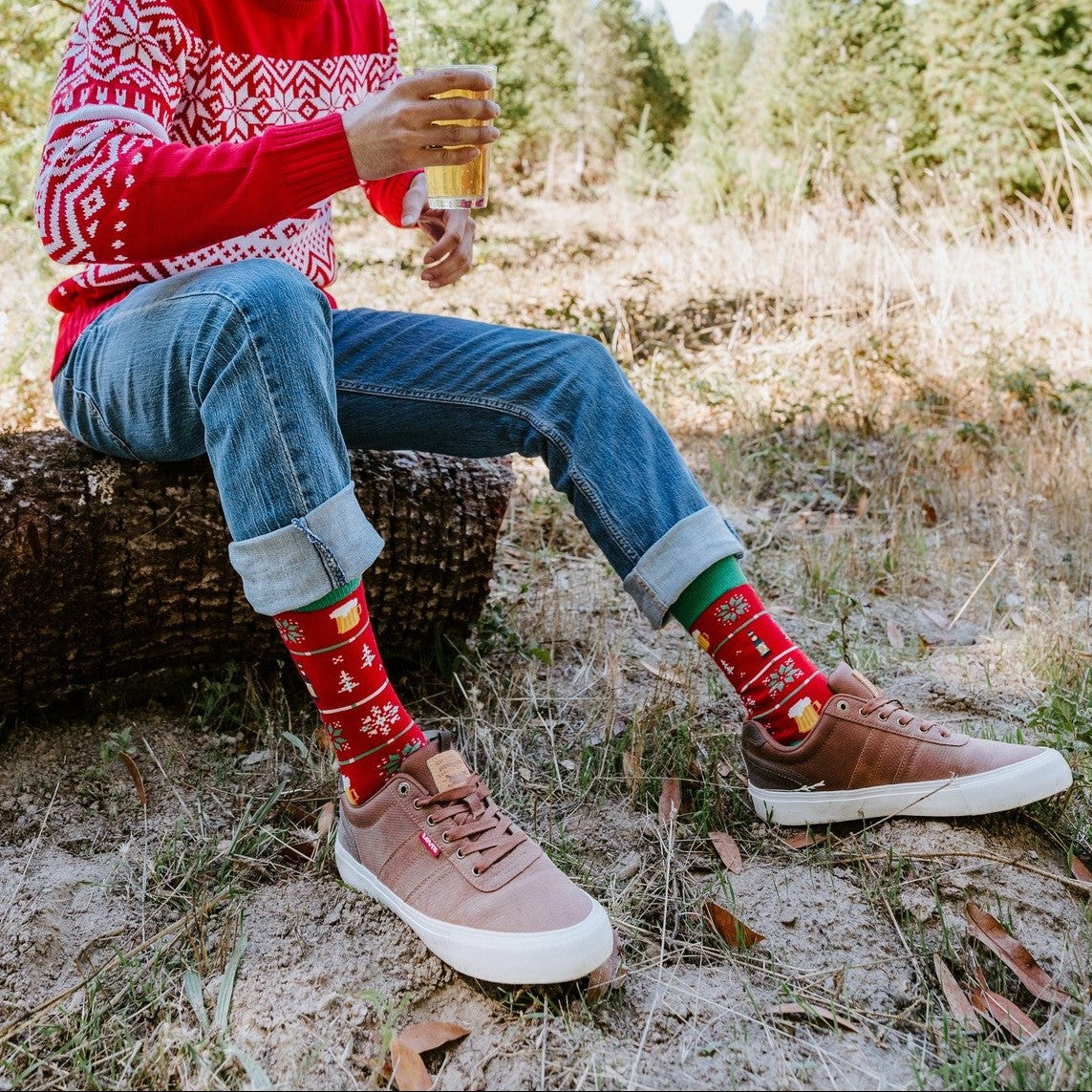 Man looks down smiling at the beer in his hand while seated on a log, jeans cuffed above Beer Sweater in Red socks and pink shoes. [lf]