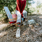 Smiling woman in a red cable knit sweater and Santa hat sits on a floral painted chair beside wrapped gifts wearing Beer Sweater in Red socks. [lf]