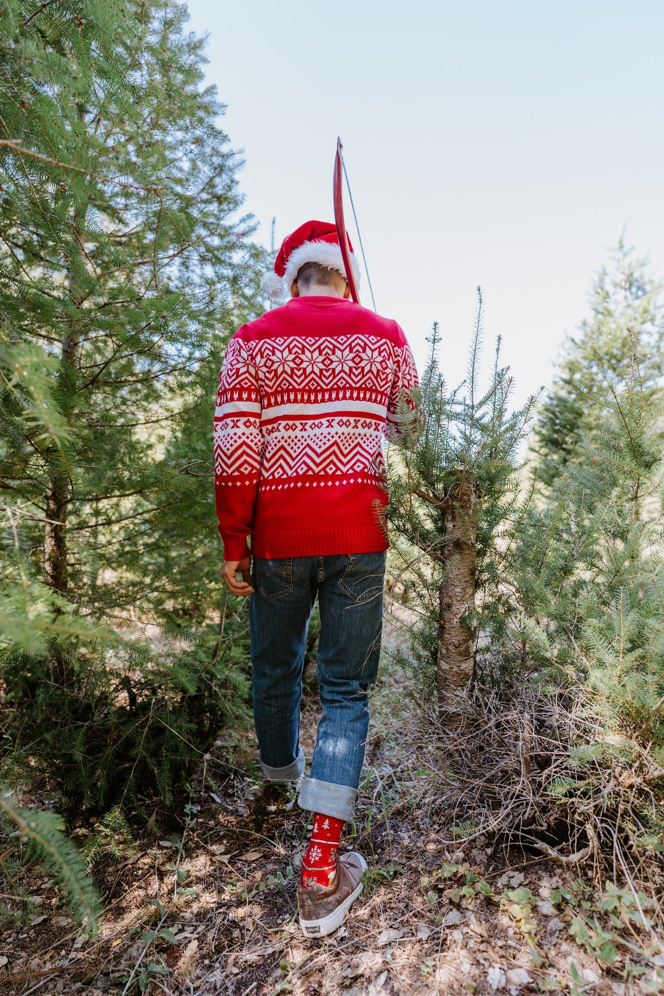 Back of a man in a red snowflake sweater walking through a Christmas tree farm with Beer Sweater in Red socks visible above his hiking boots. [lf]