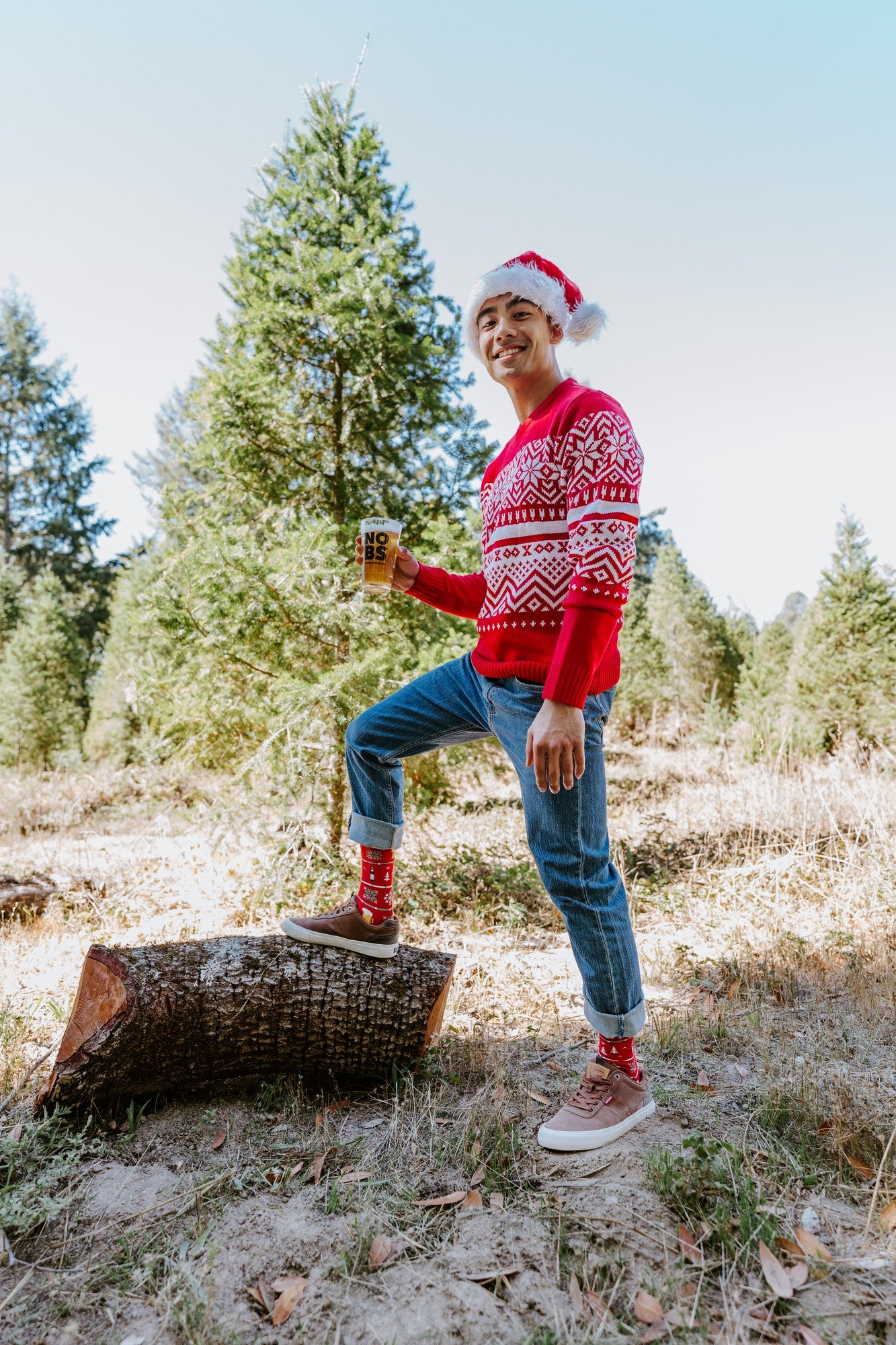 Grinning man in a Santa hat and red Nordic sweater poses with one foot on a log and a pint of beer in hand, showing his Beer Sweater in Red socks. [lf]