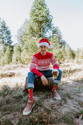 Vertical portrait of a man on a basket stool in a tree farm holding a pint glass and showing off Beer Sweater in Red socks with cuffed jeans. [lf]