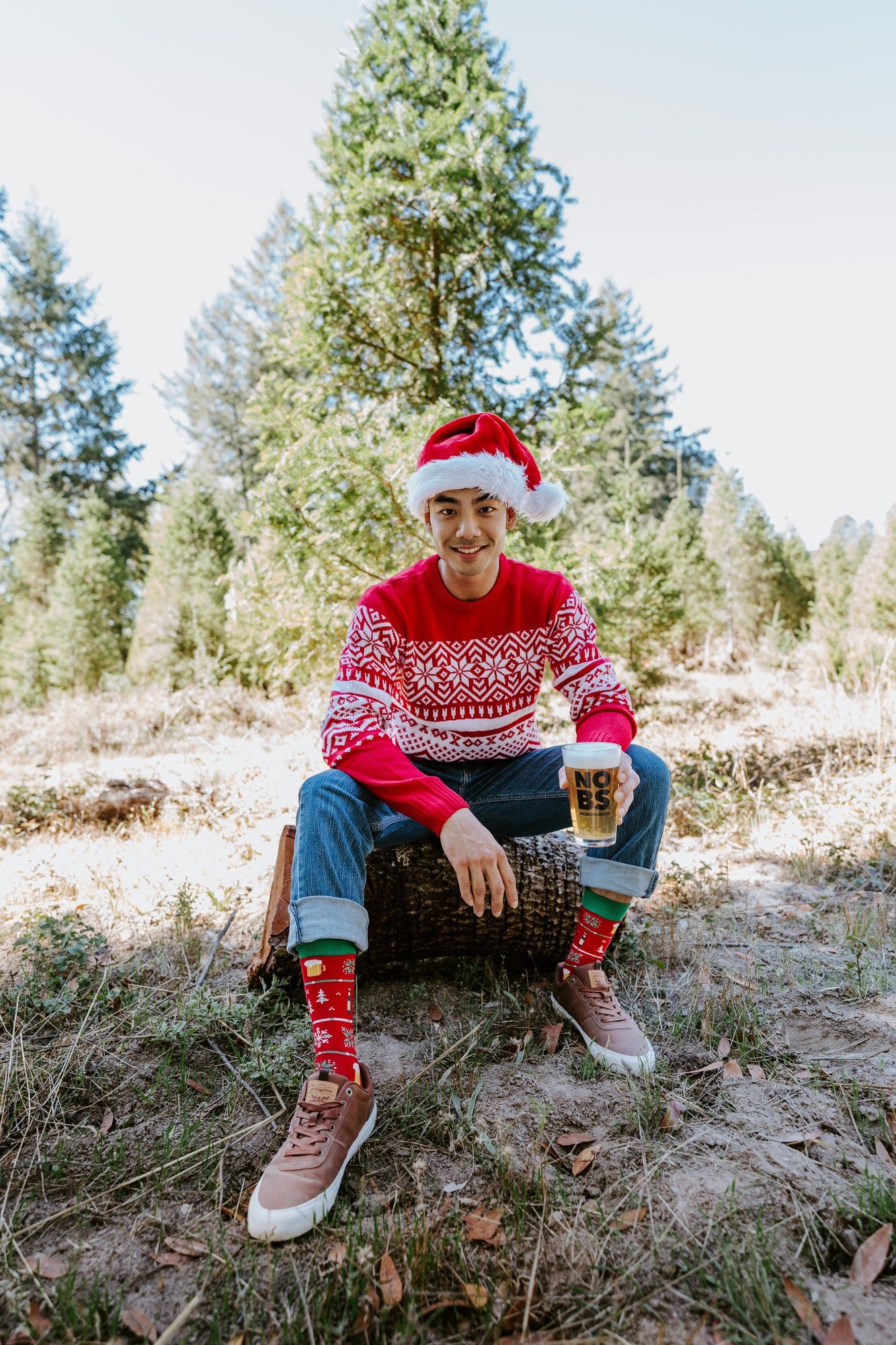 Vertical portrait of a man on a basket stool in a tree farm holding a pint glass and showing off Beer Sweater in Red socks with cuffed jeans. [lf]