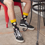Cropped view of a person eating a pastry at a metal cafe table with crossed ankles showing black Bacon X Eggs in Black socks above black skate shoes. [lf]