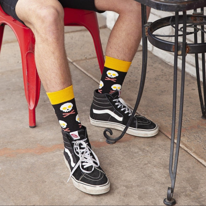 Cropped view of a person eating a pastry at a metal cafe table with crossed ankles showing black Bacon X Eggs in Black socks above black skate shoes. [lf]