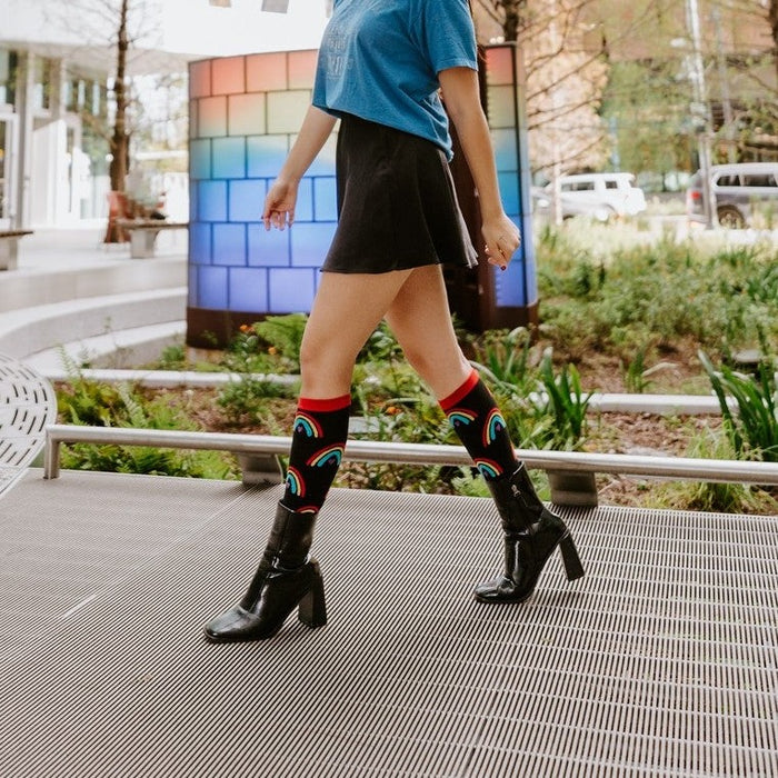 A woman walks across a textured plaza in front of a colorful mosaic wall and a stepped modern high-rise, wearing Boho Rainbow socks with red cuffs above glossy black block-heel boots. [lf]