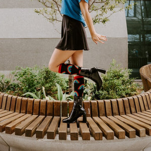 A woman balances playfully on a curved wooden bench with one arm raised, her red-cuffed Boho Rainbow socks and glossy black boots crossed at the ankles. [lf]