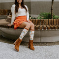 A woman sits smiling on a curved wooden bench in a planted courtyard, showing off her Boho Rainbow in Ivory Heather socks tucked into brown suede boots beneath an orange tiered skirt. [lf]
