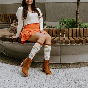 A woman sits smiling on a curved wooden bench in a planted courtyard, showing off her Boho Rainbow in Ivory Heather socks tucked into brown suede boots beneath an orange tiered skirt. [lf]