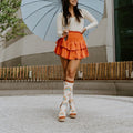 A woman poses mid-stride with one heel kicked up under a soft blue umbrella, her Boho Rainbow in Ivory Heather socks paired with strappy cream platform sandals and an orange ruffled skirt. [lf]