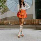 A woman poses mid-stride with one heel kicked up under a soft blue umbrella, her Boho Rainbow in Ivory Heather socks paired with strappy cream platform sandals and an orange ruffled skirt. [lf]