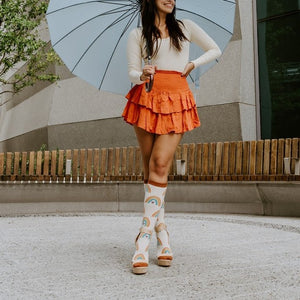 A woman poses mid-stride with one heel kicked up under a soft blue umbrella, her Boho Rainbow in Ivory Heather socks paired with strappy cream platform sandals and an orange ruffled skirt. [lf]