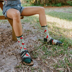 Woman seated on a wooden picnic bench under a black umbrella, lifting binoculars to her eyes while her Cardinal Tracks socks in Mint with red bird motifs peek above sport sandals. [lf]