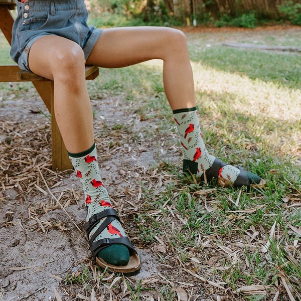 Woman seated on a wooden picnic bench under a black umbrella, lifting binoculars to her eyes while her Cardinal Tracks socks in Mint with red bird motifs peek above sport sandals. [lf]