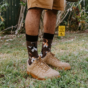 Standing on a grassy backyard lawn in tan work shorts and tan boots, a person holds a tied poop bag and leash while wearing Cat Butts socks in Brown Heather covered in playful kitty rear prints. [lf]