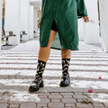 A smiling woman in a green graduation gown holds a champagne bottle under a flowering white pergola, wearing Champagne Toast in Black socks with chunky black boots. [lf]