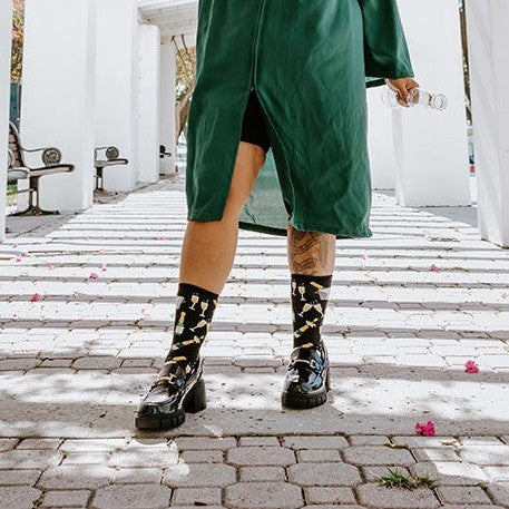 A smiling woman in a green graduation gown holds a champagne bottle under a flowering white pergola, wearing Champagne Toast in Black socks with chunky black boots. [lf]