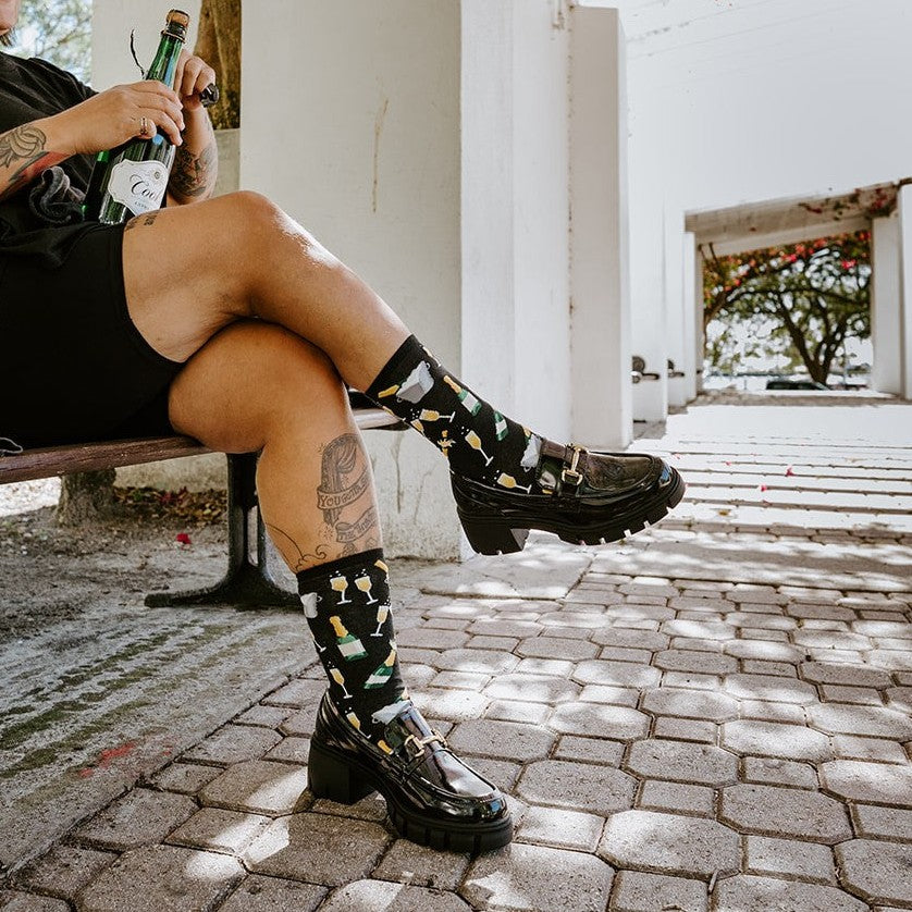 Seated on a park bench with crossed legs and a flute in hand, she sips from a green bottle showing off her Champagne Toast in Black socks. [lf]