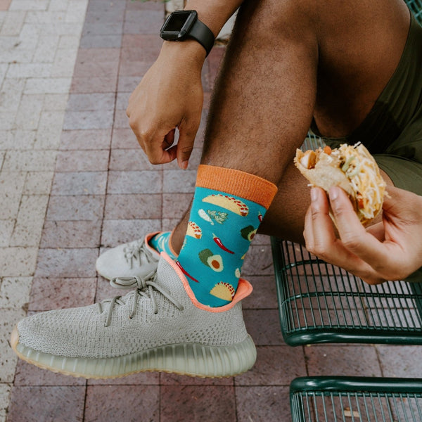 A tight close-up frames one ankle, a taco-filled hand, and a smartwatch above Something To Taco Bout Bamboo in Blue socks on a brick walkway. [lf]