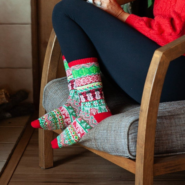Woman in a red knit sweater laughs while holding a mug beneath a leafy houseplant, with Tis the Season Sweater in Red and Green socks peeking out below her leggings. [lf]