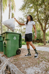 Vertical shot of the same curbside moment with palms and trees in the background as he lifts the trash bag, Trash Bandit Bamboo socks in Green peeking out between his shorts and sneakers. [lf]
