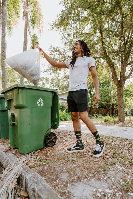 Vertical shot of the same curbside moment with palms and trees in the background as he lifts the trash bag, Trash Bandit Bamboo socks in Green peeking out between his shorts and sneakers. [lf]