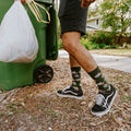 Side view as he grips the bin handle with a full trash bag in the other hand, the raccoon and tree print on his Trash Bandit Bamboo socks in Green sharp in the late afternoon light. [lf]