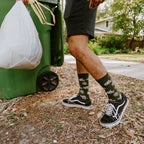 Side view as he grips the bin handle with a full trash bag in the other hand, the raccoon and tree print on his Trash Bandit Bamboo socks in Green sharp in the late afternoon light. [lf]
