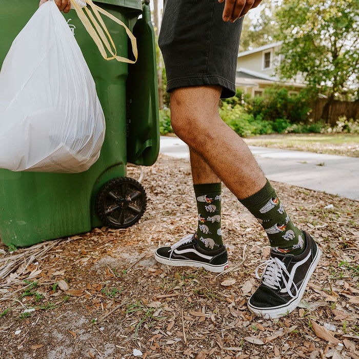 Side view as he grips the bin handle with a full trash bag in the other hand, the raccoon and tree print on his Trash Bandit Bamboo socks in Green sharp in the late afternoon light. [lf]