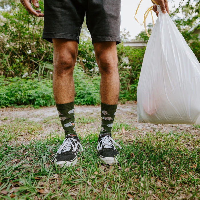 Both feet planted in the grass with a white trash bag swinging at his side, showing the full ribbed cuff and raccoon motif of Trash Bandit Bamboo socks in Green. [lf]