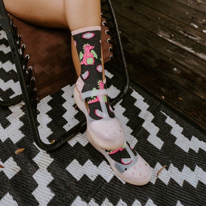 A vertical portrait of a woman reclining outdoors in a pink romper, sipping wine with crossed ankles in Dragonfruit in Charcoal Heather socks. [lf]