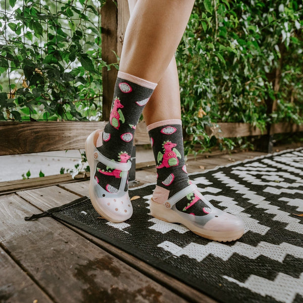 A tight crop on crossed ankles in Dragonfruit in Charcoal Heather socks featuring pink fruit motifs against the black-and-white patio rug. [lf]
