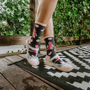 A tight crop on crossed ankles in Dragonfruit in Charcoal Heather socks featuring pink fruit motifs against the black-and-white patio rug. [lf]