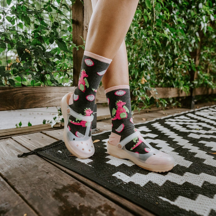 A tight crop on crossed ankles in Dragonfruit in Charcoal Heather socks featuring pink fruit motifs against the black-and-white patio rug. [lf]