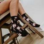 Top down view of crossed legs on a wooden chair, with Fine Like Boxed Wine socks in Black showcasing rows of pink boxed-wine cartons and stemmed glasses while a hand cradles a red wine pour. [lf]
