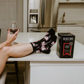 Smirking at the camera with feet propped on a white kitchen island next to a Black Box cabernet carton, a woman models Fine Like Boxed Wine socks in Black covered in pink wine box and glass illustrations. [lf]