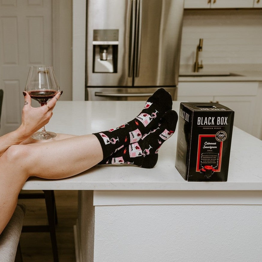 Smirking at the camera with feet propped on a white kitchen island next to a Black Box cabernet carton, a woman models Fine Like Boxed Wine socks in Black covered in pink wine box and glass illustrations. [lf]
