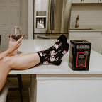 Smirking at the camera with feet propped on a white kitchen island next to a Black Box cabernet carton, a woman models Fine Like Boxed Wine socks in Black covered in pink wine box and glass illustrations. [lf]