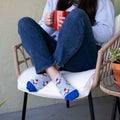 A woman curls up in a wicker chair with a red mug, her crossed feet showing Growing Gnomes ankle socks in heather gray dotted with tiny gnomes and stars, paired with cuffed jeans on a sunny patio. [lf]