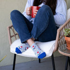 A woman curls up in a wicker chair with a red mug, her crossed feet showing Growing Gnomes ankle socks in heather gray dotted with tiny gnomes and stars, paired with cuffed jeans on a sunny patio. [lf]